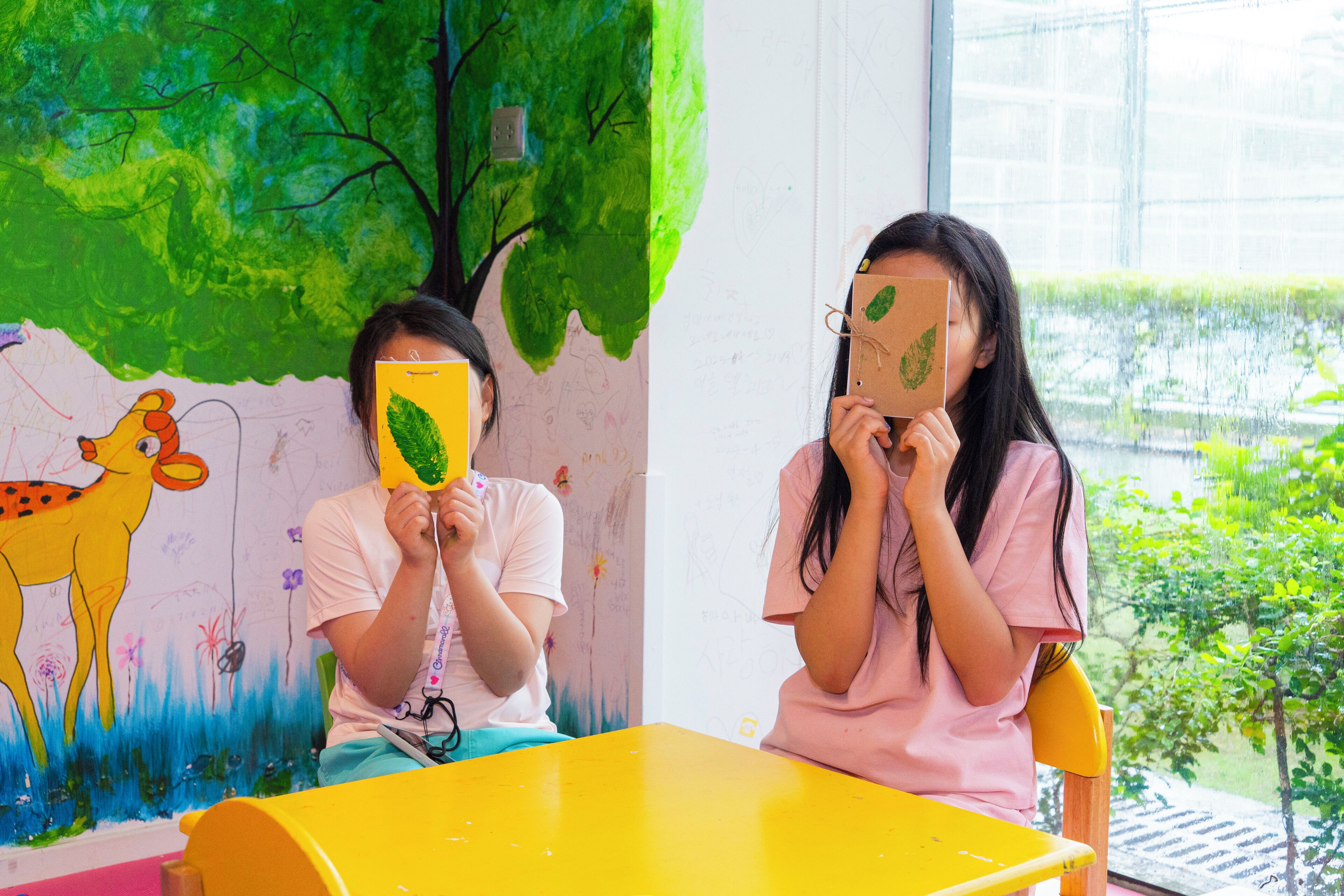 Two children sitting at a yellow table in a room with a green mural and a window with a view of greenery.