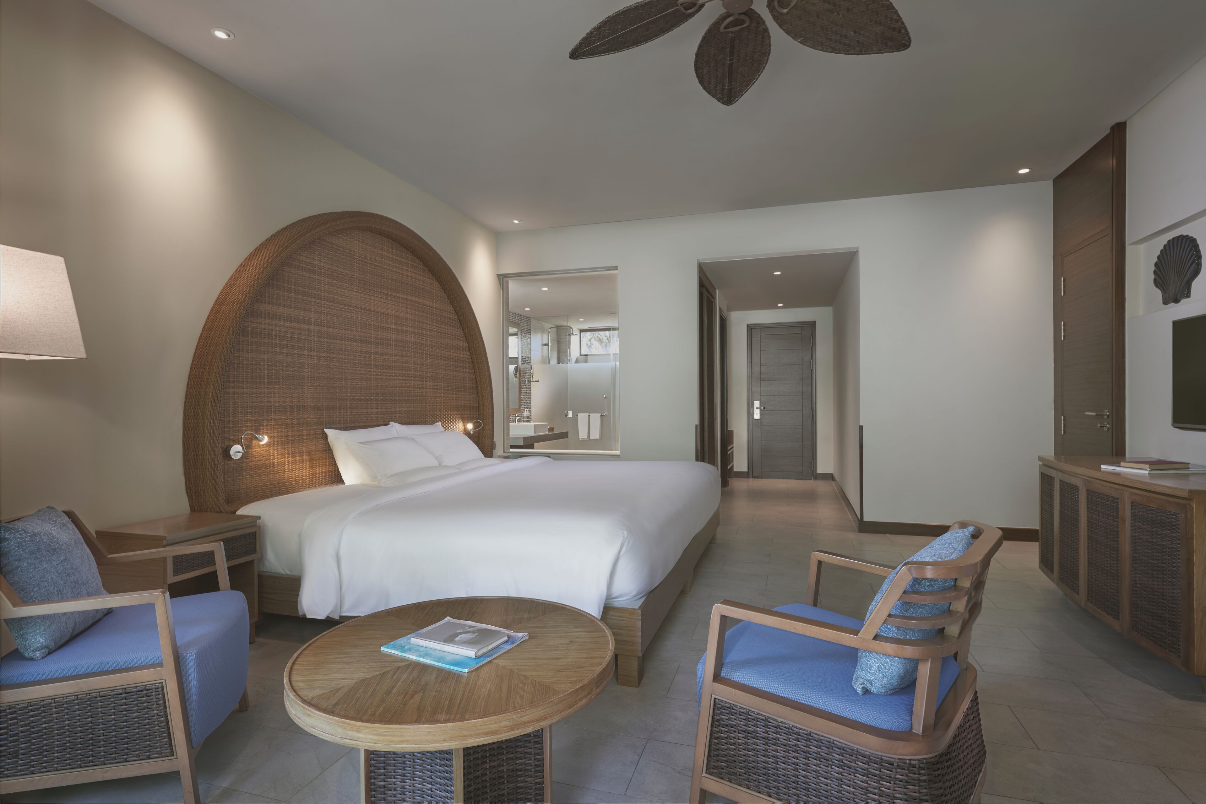 A hotel room with a large bed, two chairs, a round table, a ceiling fan, and a TV on a cabinet.