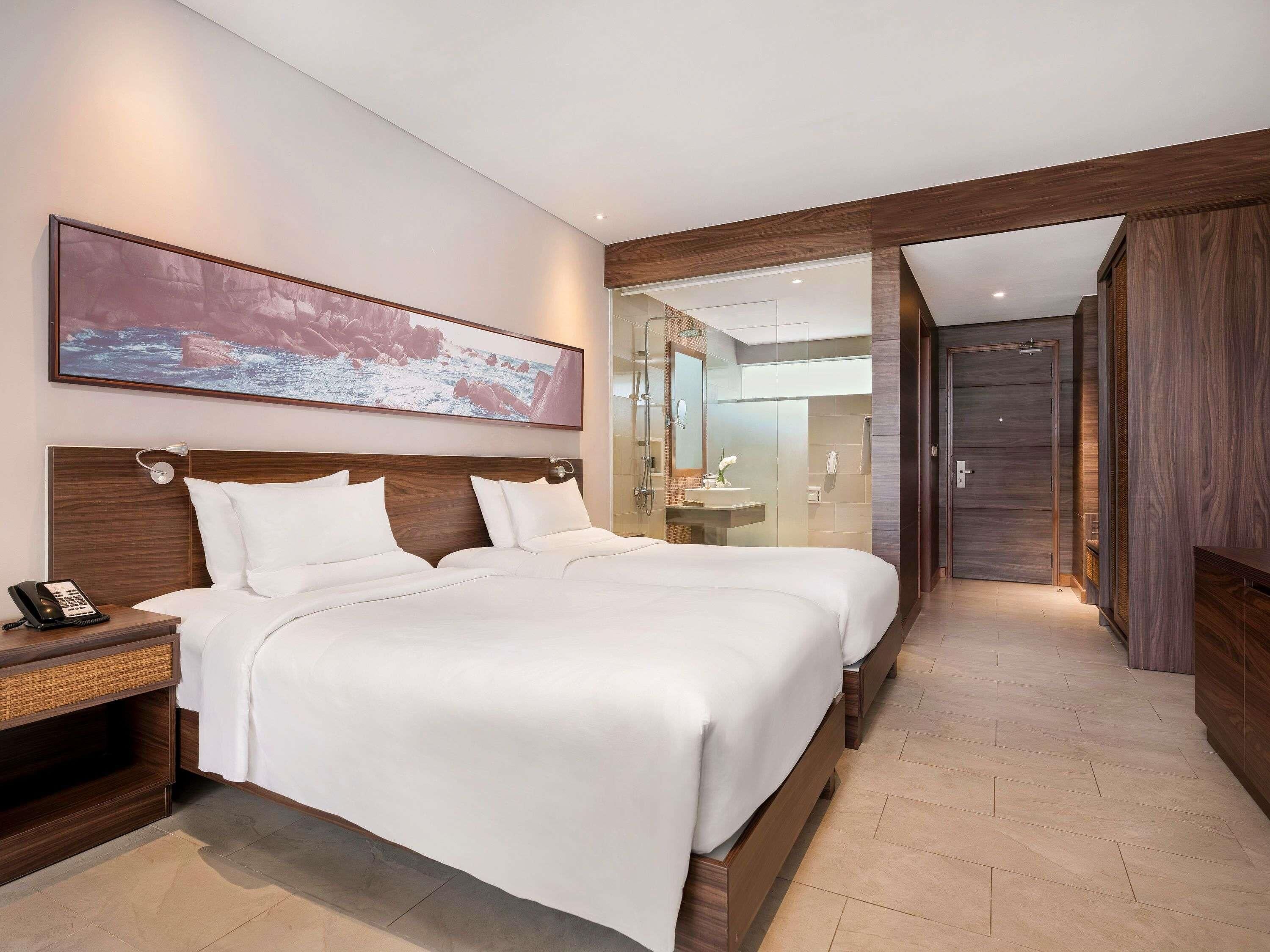 A hotel room with two beds, a large painting, a phone on the nightstand, and a bathroom visible through a glass partition.