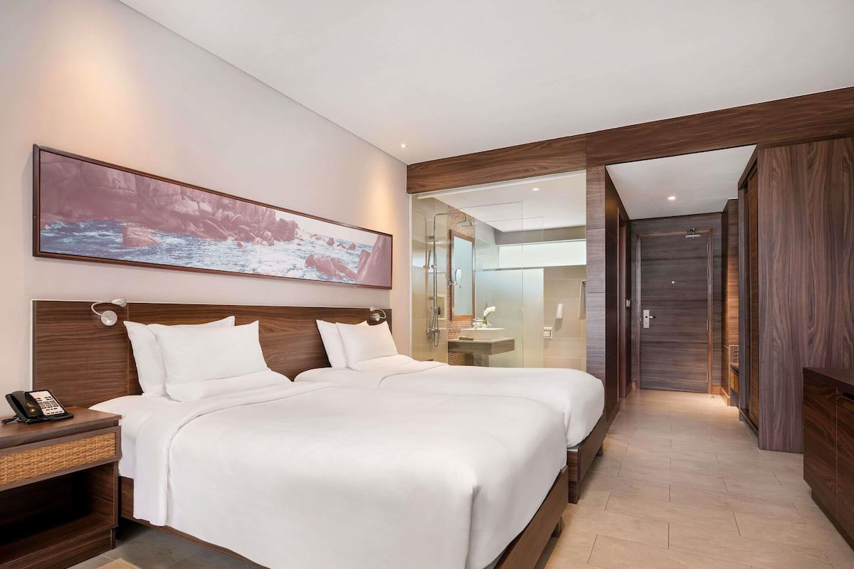 A hotel room with two beds, a large painting, a phone on the nightstand, and a bathroom visible through a glass partition.