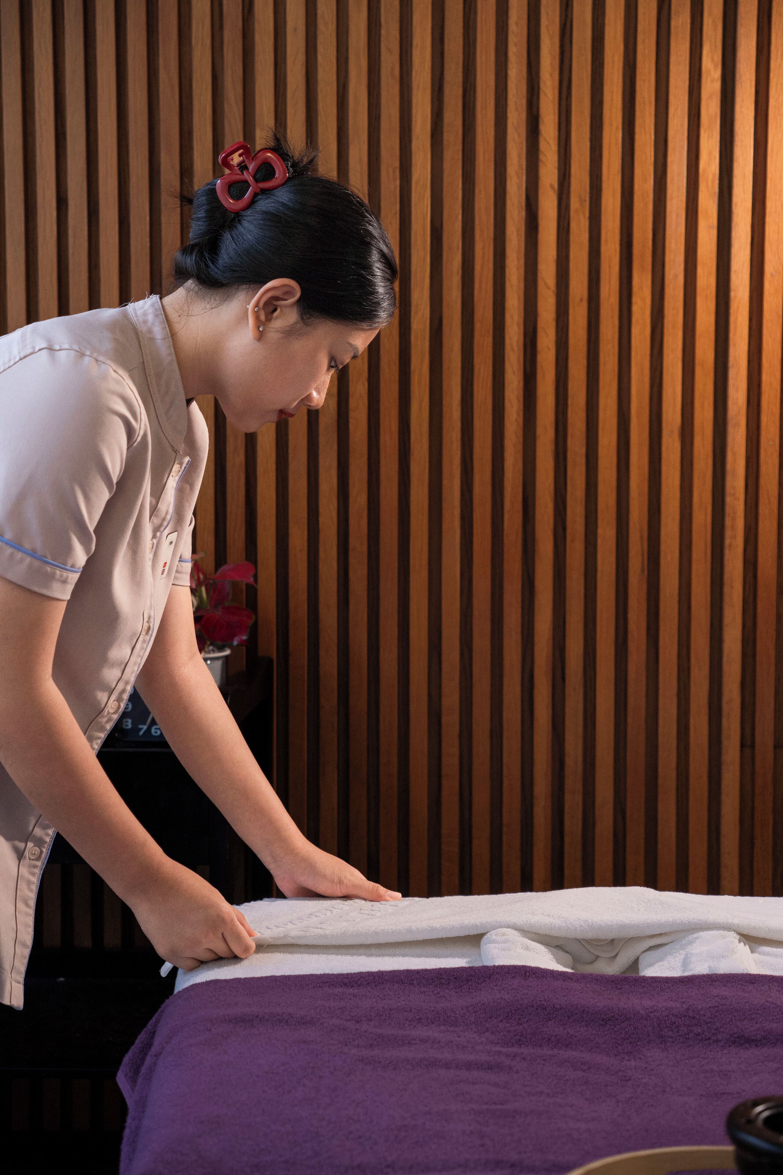 A person attending to a bed with a purple blanket, wooden slat wall, and a small vase with flowers in the background.
