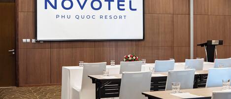 Een vergaderzaal bij Novotel met een groot scherm waarop de hotelnaam en -locatie staan, geordende tafels met stoelen en een spreekgestoelte.