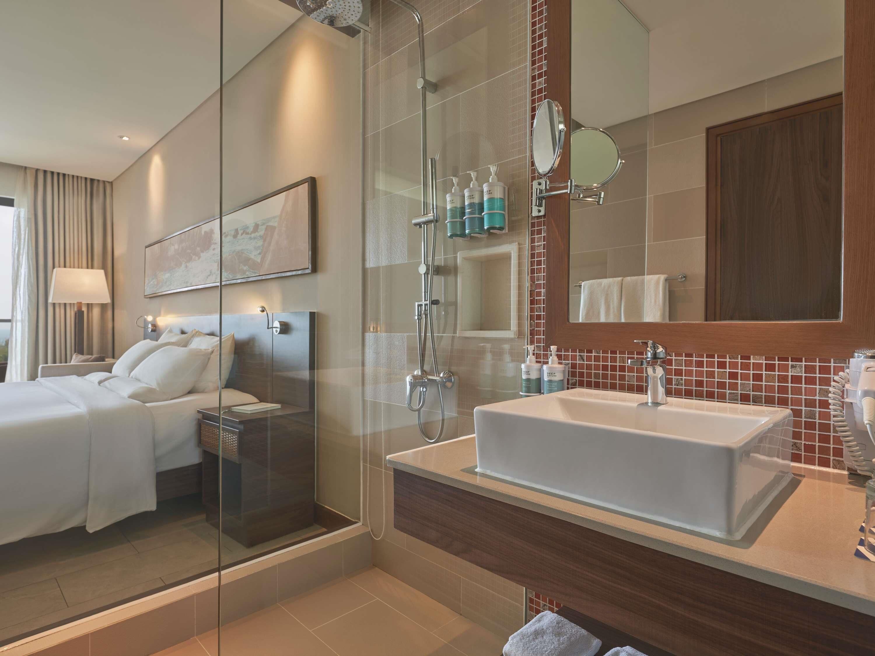 Een moderne hotelkamer met een glazen douchecabine, een groot bed, een nachtkastje, een vastgeplakt spiegel en een badkamer met een wastafel en toiletartikelen.