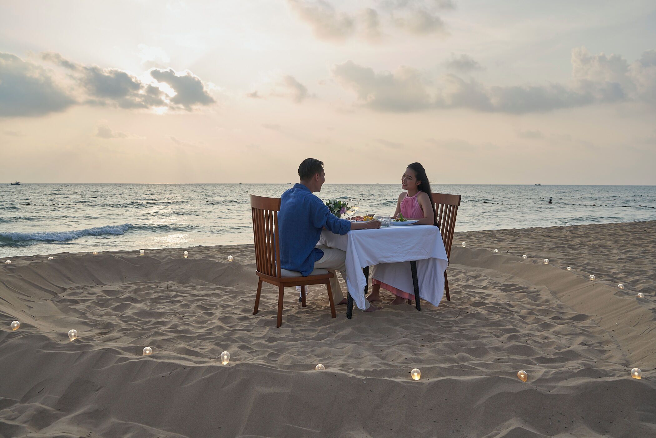 Una pareja cenando en una mesa sobre una playa arenosa, con el océano de fondo.
