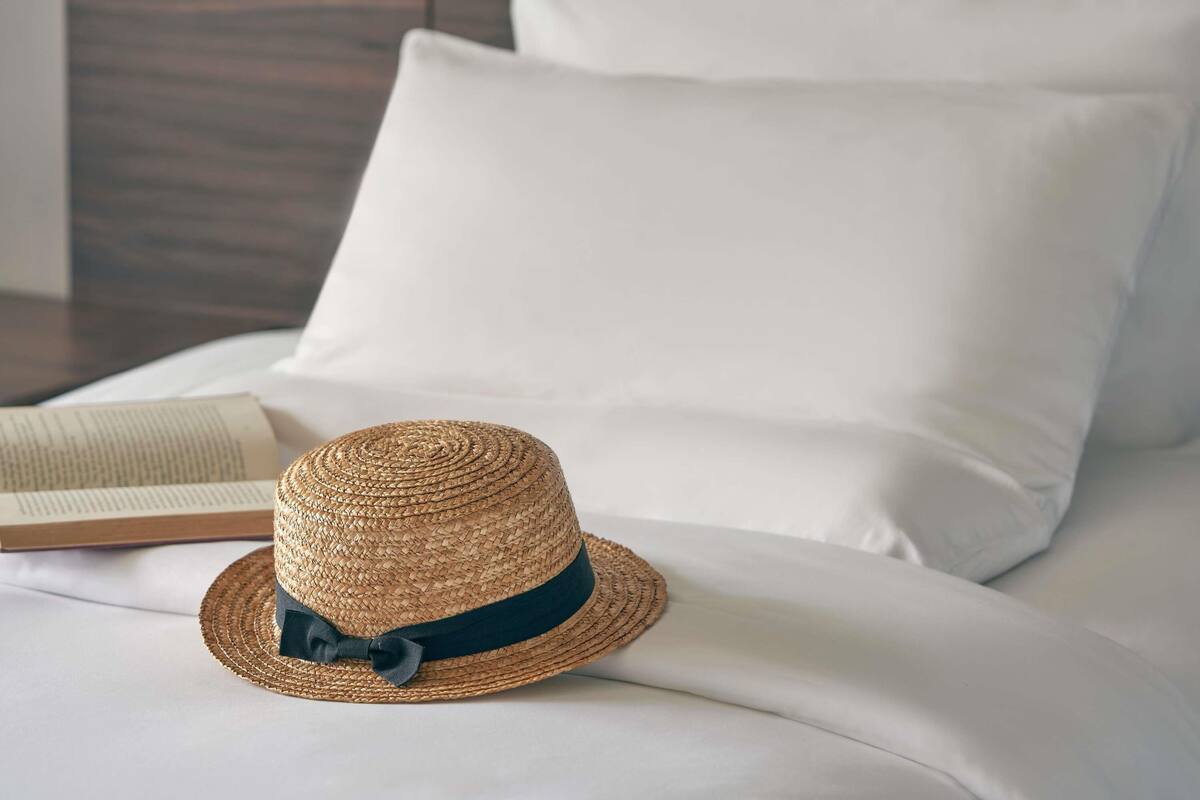 A straw hat with a dark ribbon on a bed with white sheets and pillows.