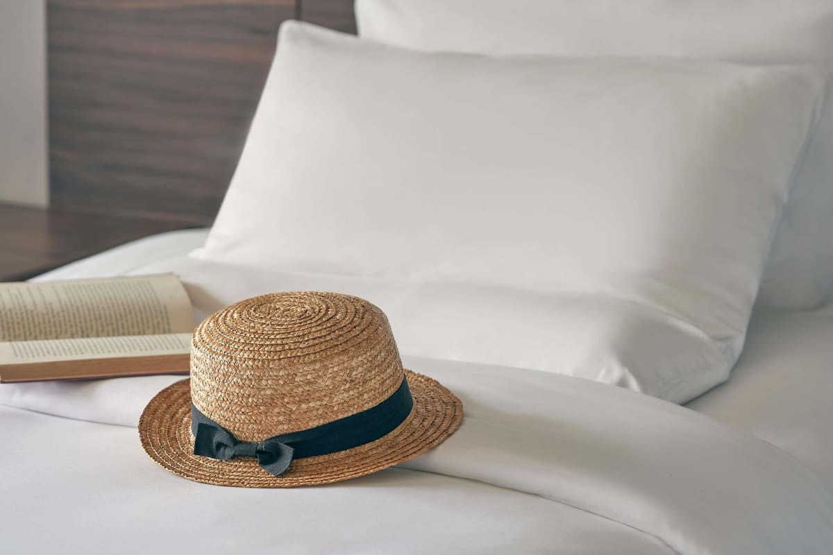 A straw hat with a dark ribbon on a bed with white sheets and pillows.