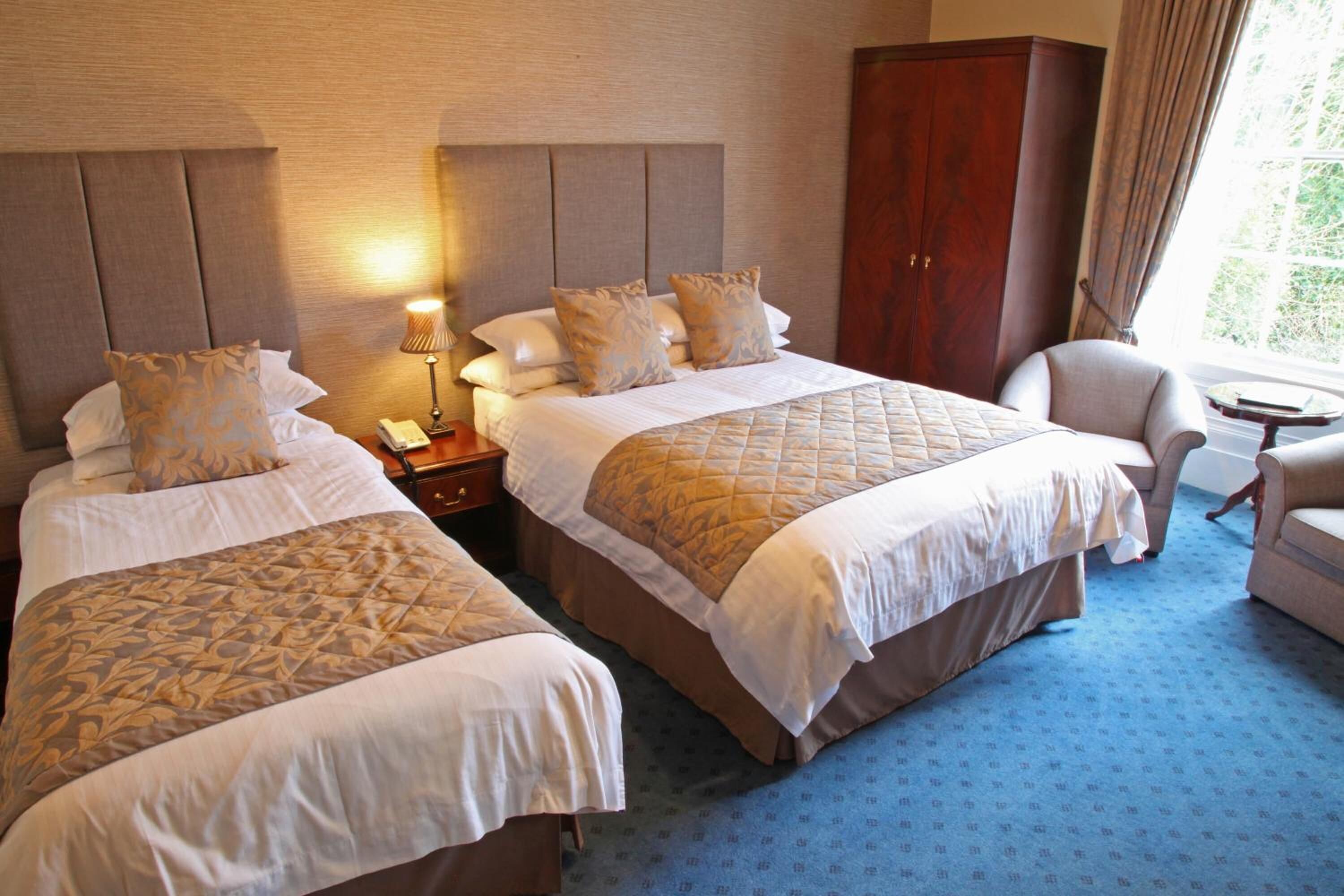 A hotel room with two beds, a wardrobe, a chair, a small table, and a lamp.
