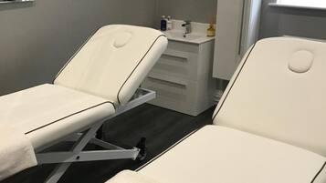 A clinical examination room with two examination beds, a white cabinet, and a window with blinds.