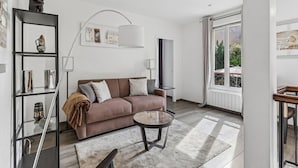 A modern living room with a sofa, a coffee table, a bookshelf, and a large window. - Loft Fraternité (Bagnolet)