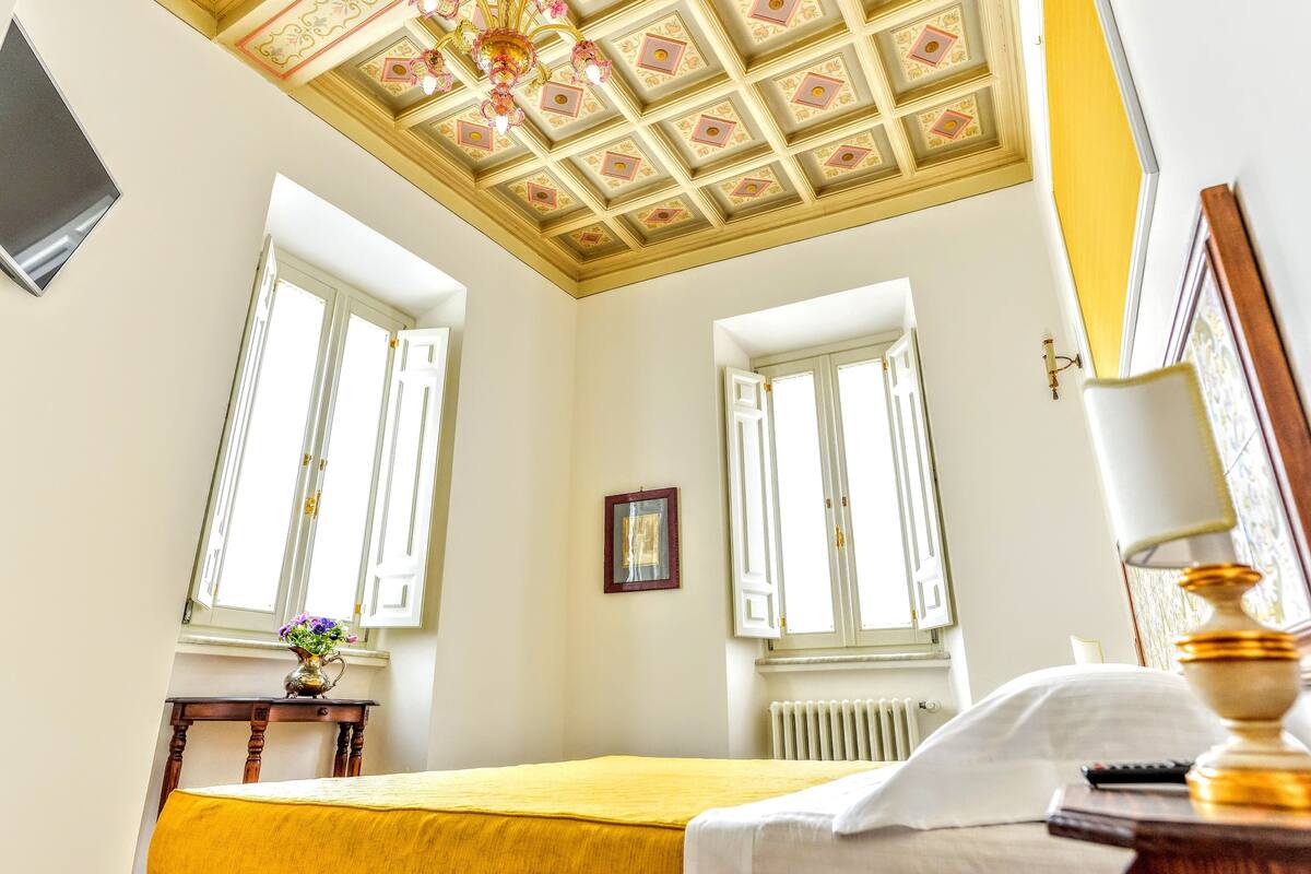 Una habitación de hotel con cama, ventana, radiador, marco de cuadro y candelabro.