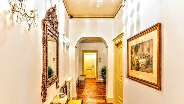 A hallway with a wooden floor, a table with a yellow cushion, a mirror, and a framed painting on the wall. - Arpinelli Relais (Rome)