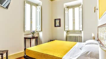 Een slaapkamer met een bed, twee ramen, een radiator, een tafeltje met een vaas en een afbeelding aan de muur.