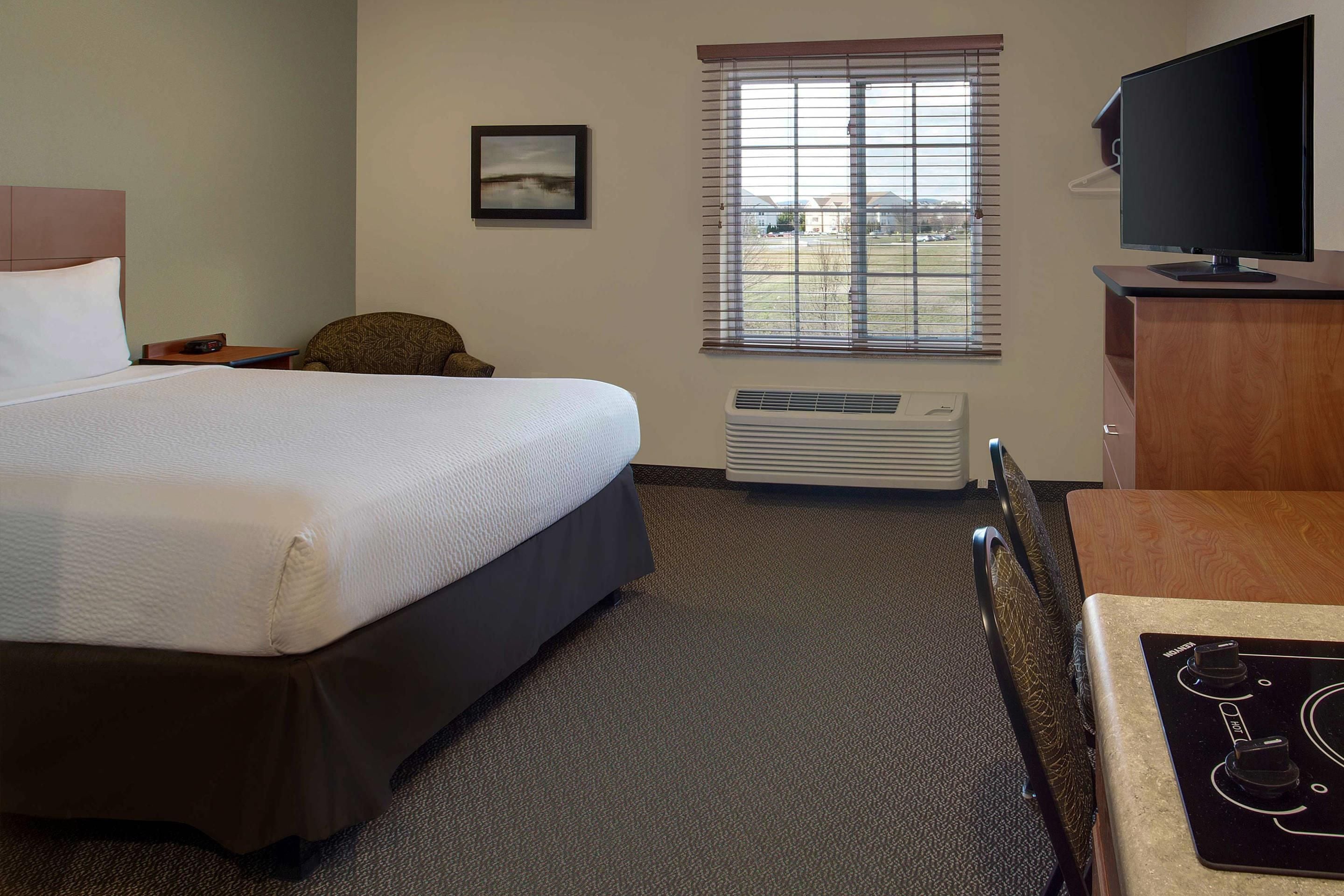 A hotel room with a bed, a chair, a desk with a computer, a television, a window with blinds, and a picture on the wall.
