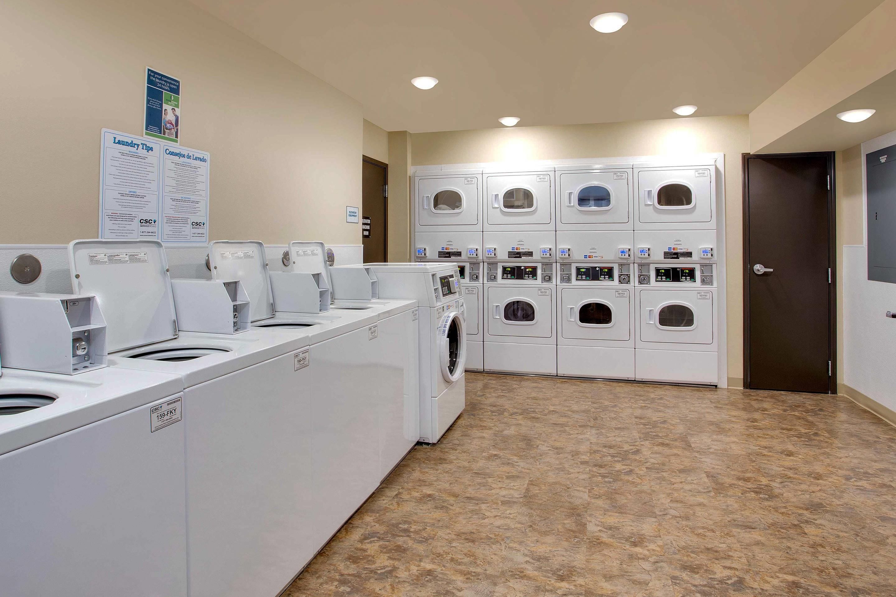 A laundry facility with multiple washing machines and dryers, a sign with laundry instructions, and a door to the right.