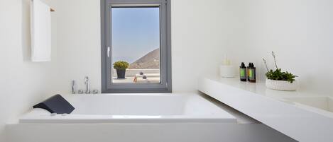 A modern bathroom with a large bathtub, a window with a view of a mountainous landscape, a towel rack, and a shelf with toiletries.