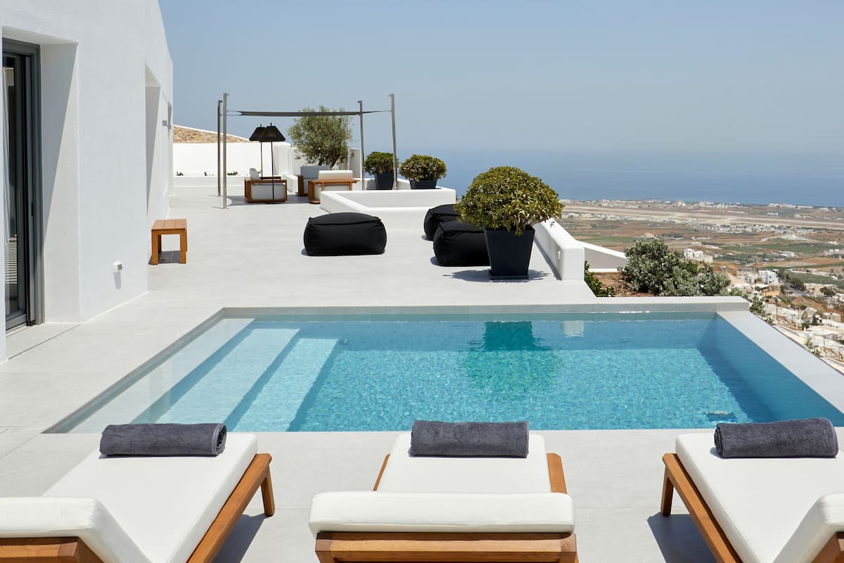 A rooftop infinity pool with lounge chairs and potted plants, overlooking a coastal landscape.