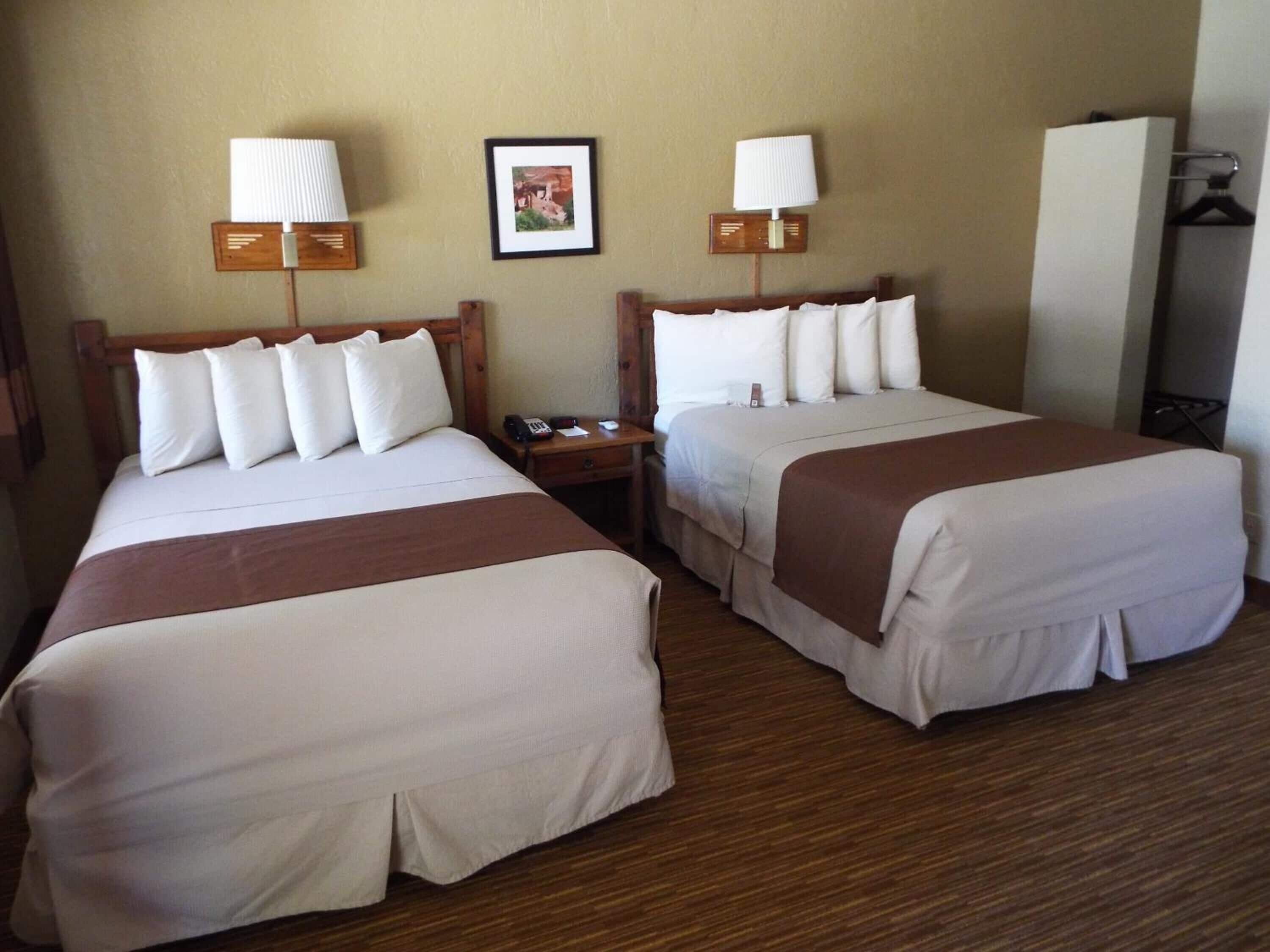 A hotel room with two beds, each with white bedding and brown headboards. There are bedside tables with lamps and a framed picture on the wall.