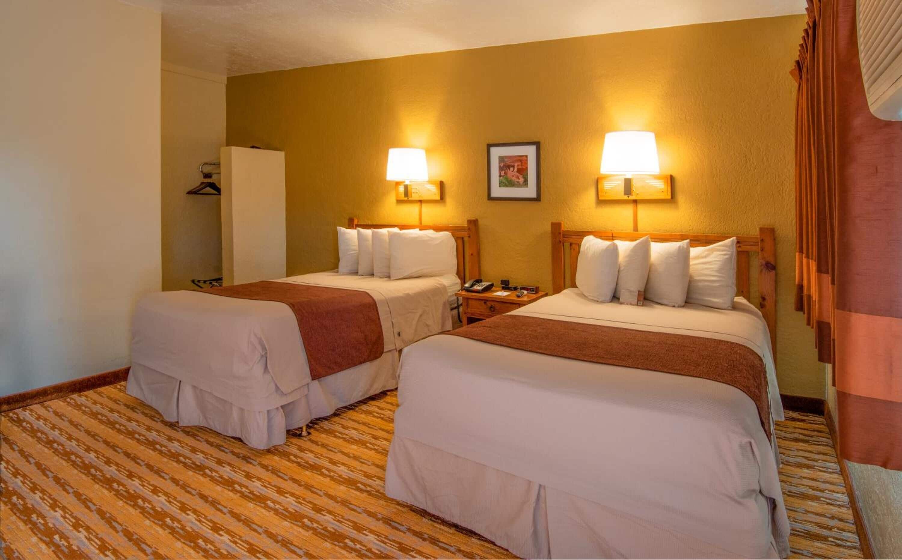Quarto de hotel com duas camas, cada uma com lençóis brancos e mantas marrons. Há duas mesas de cabeceira com abajures, um quadro emoldurado na parede e piso de madeira.