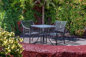 A metal table and two chairs in an outdoor patio area with a red brick wall and green foliage in the background. - Thunderbird Lodge (Chinle)