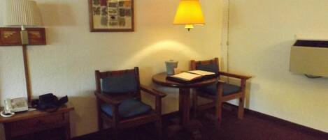 A small room with two wooden chairs, a small table, a lamp, a framed picture on the wall, and a telephone on a side table.