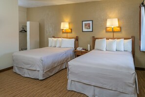 A hotel room with two beds, a bedside table with a lamp, a framed picture on the wall, and a coat rack. - Thunderbird Lodge (Chinle)