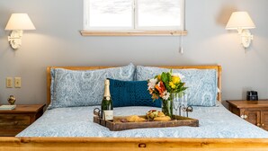 Una cama bien hecha con un edredón azul estampado, un cabecero de madera y una mesita de noche con una lámpara. En la cama hay una bandeja con una botella de champán, pasteles y un jarrón con flores.