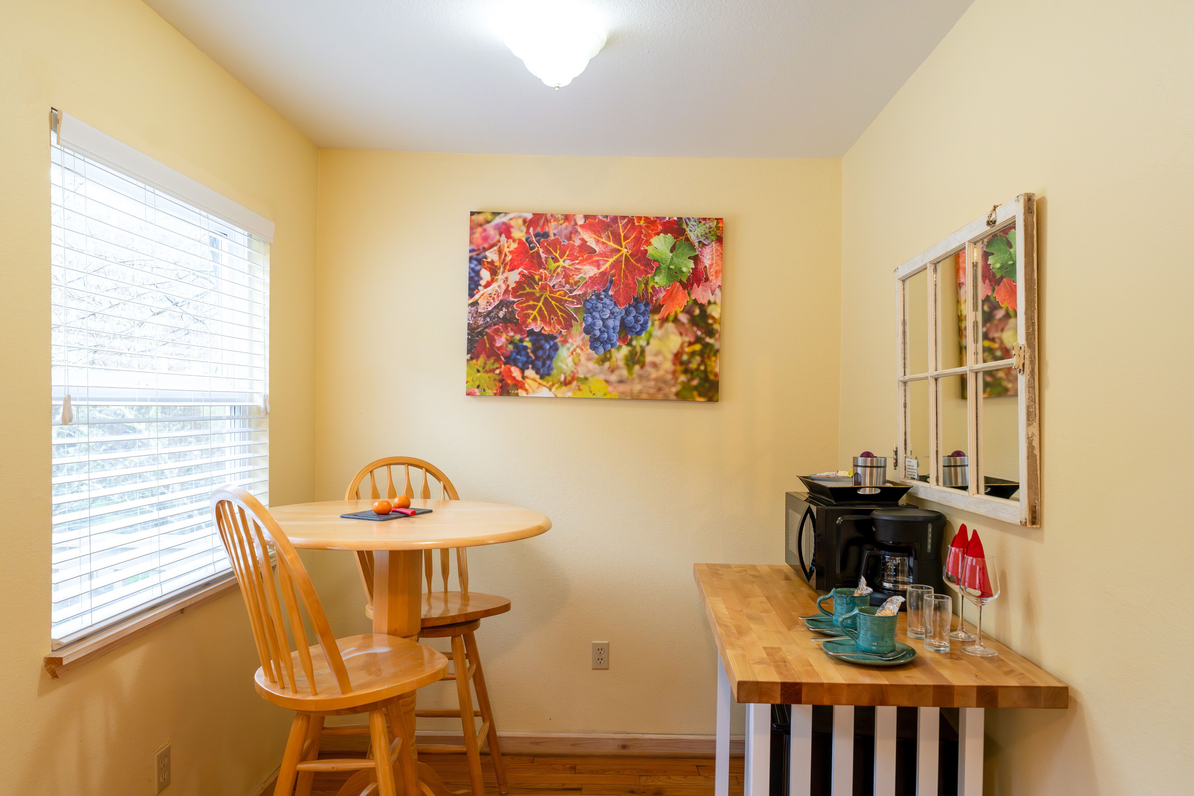 Un pequeño comedor con una mesa y sillas, una ventana con persianas, una obra de arte colorida en la pared, una cafetera y un espejo en la pared.