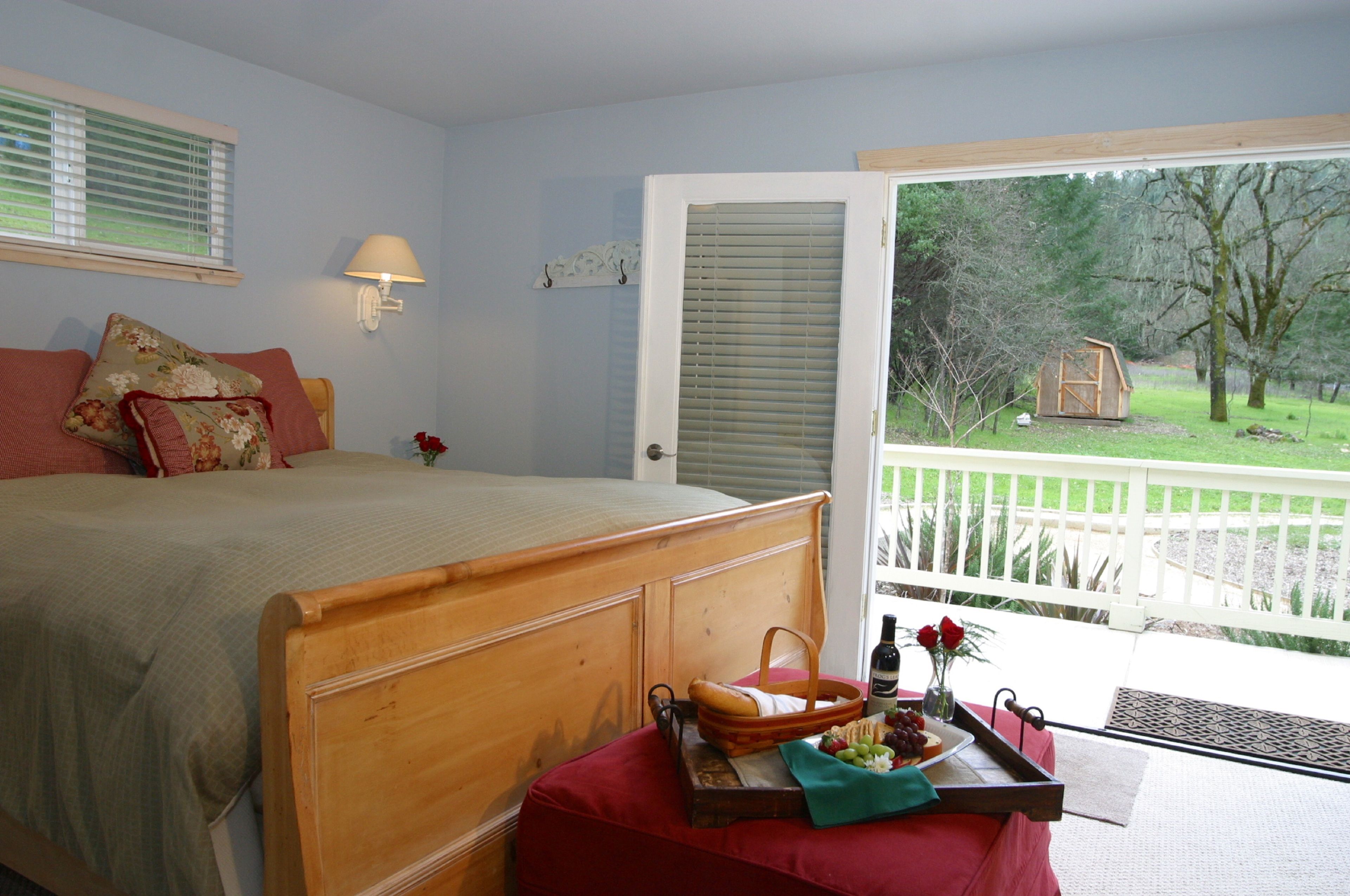 A bedroom with a wooden bed, a window with blinds, a nightstand with a tray of food, and a balcony with a view of the outdoors.