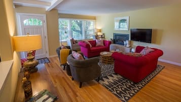 A living room with a red sofa, a grey armchair, and a patterned rug. There is a lamp on a side table, a television on the wall, and a door leading outside.