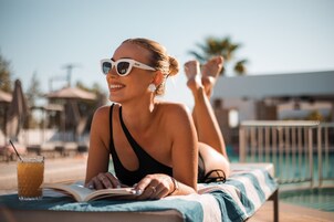 Una persona recostada en una silla de descanso junto a una piscina, con un libro y una bebida.