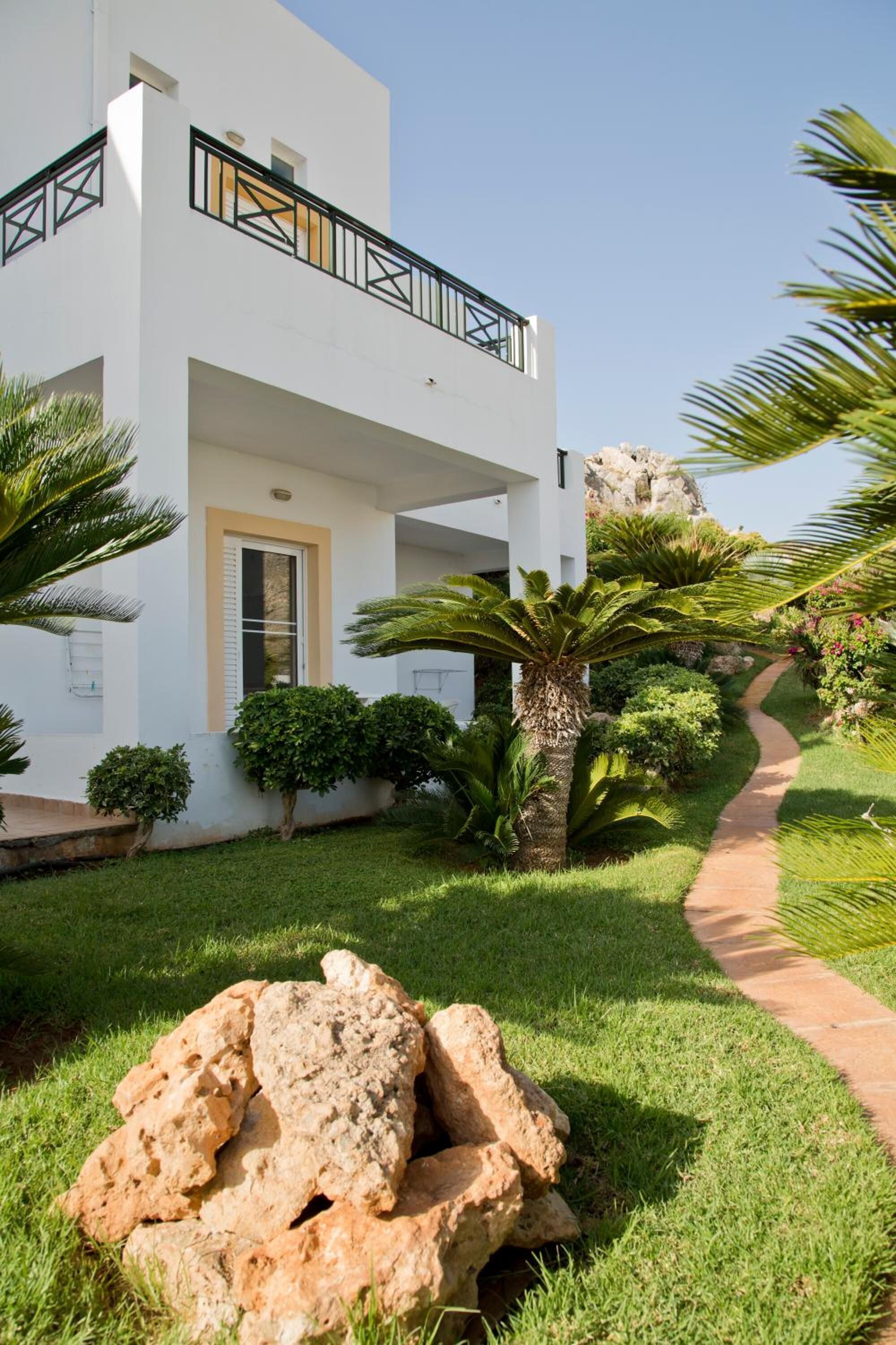 A modern white building with a balcony, surrounded by a well-maintained garden with palm trees and a stone pathway.