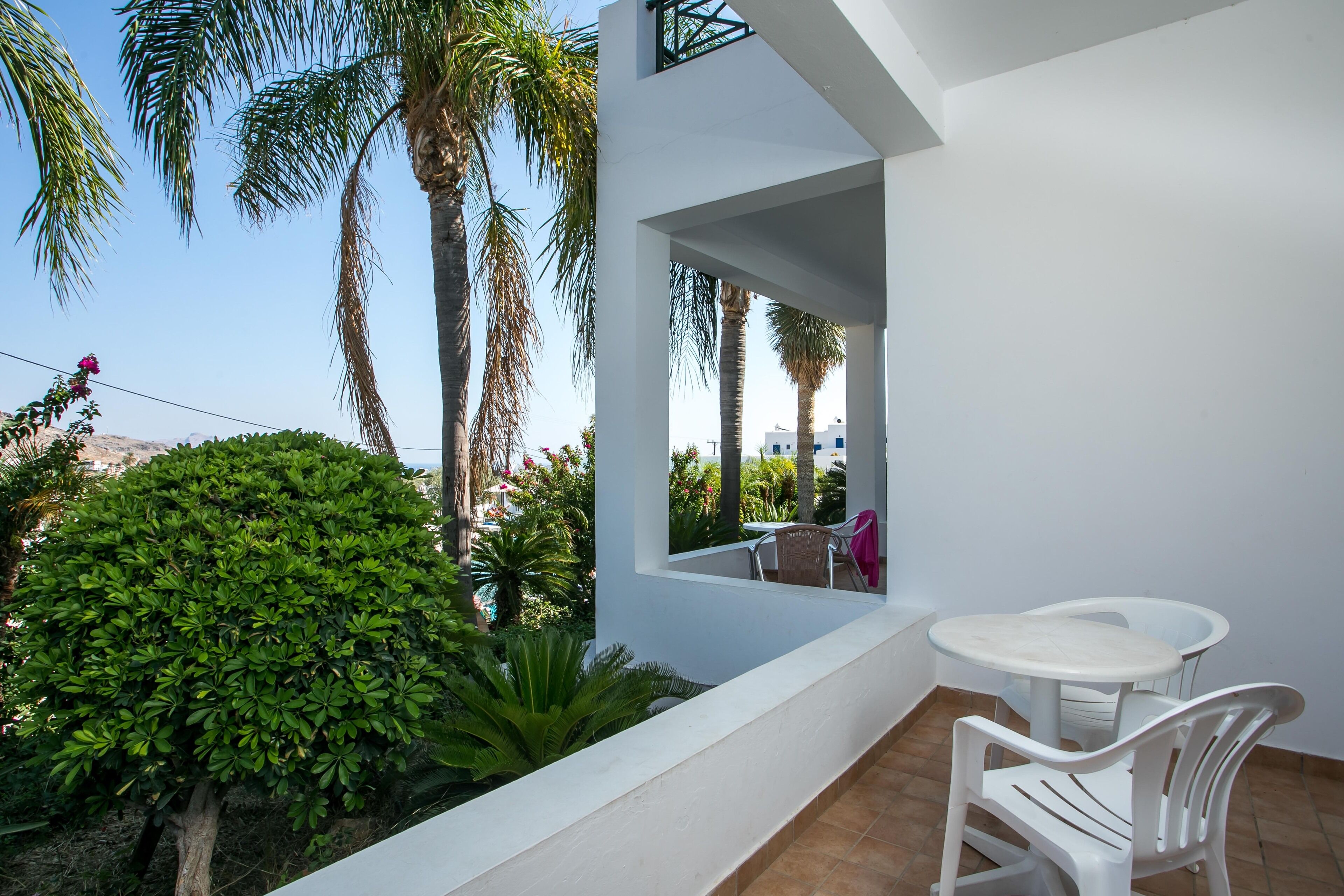 A balcony with a white table and chairs, surrounded by greenery and palm trees.