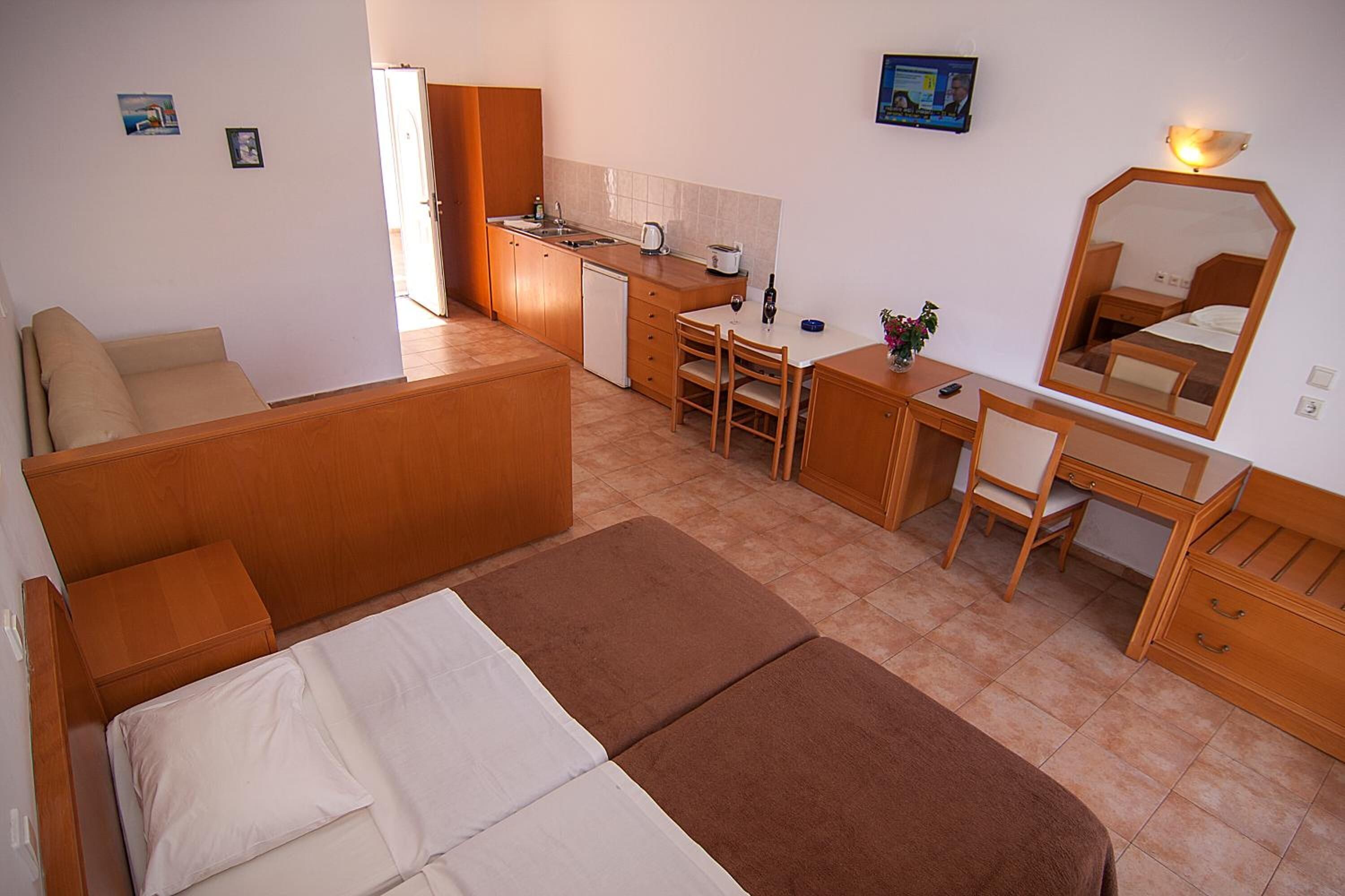 A compact hotel room with a bed, a sofa, a desk with a chair, a small kitchenette, and a mirror.