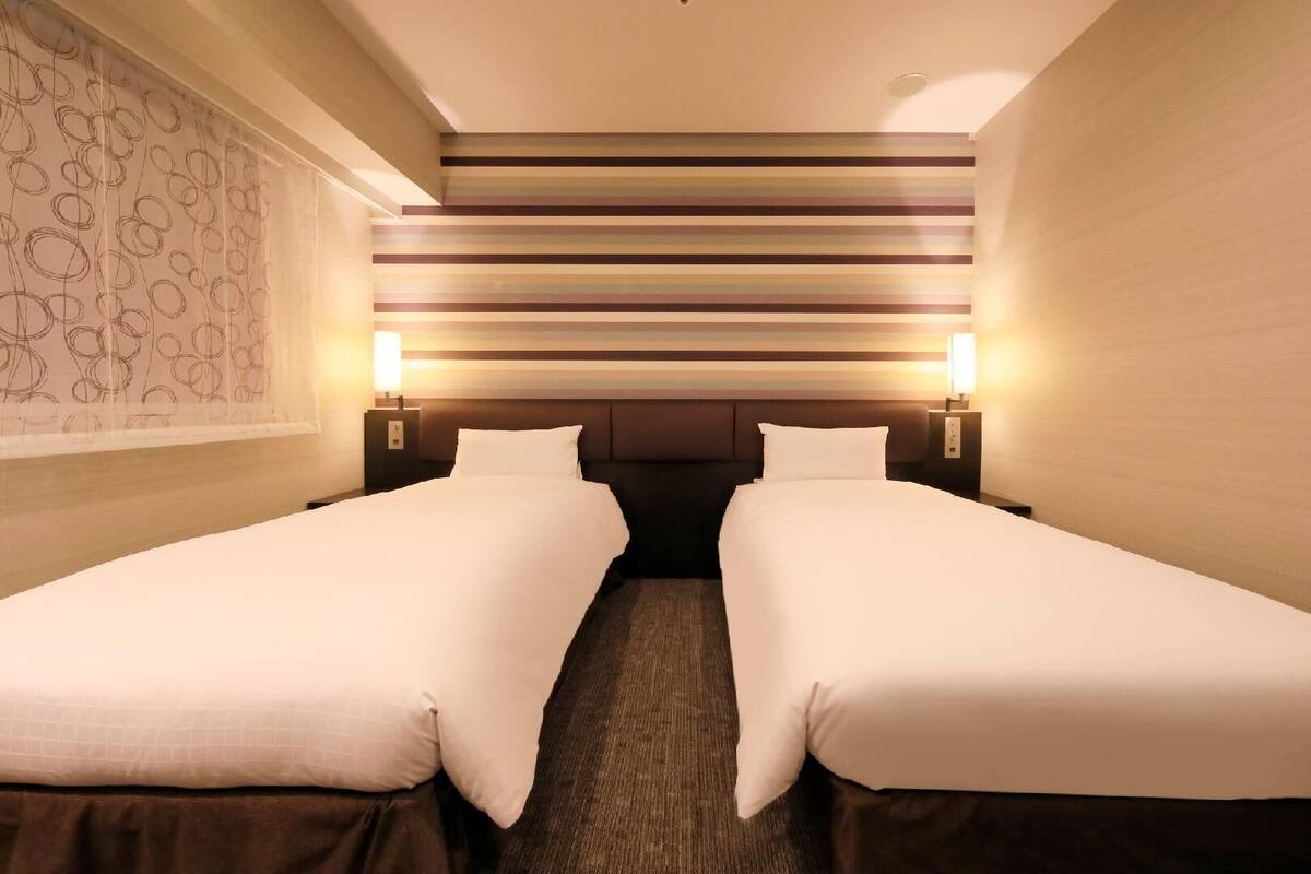 Ein Hotelzimmer mit zwei Einzelbetten, einem Kopfteil mit horizontalen Streifen und zwei Nachttischlampen.