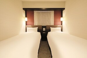 Habitació d'hotel amb dos llits, una tauleta de nit, una làmpada i una finestra amb cortina estampada.