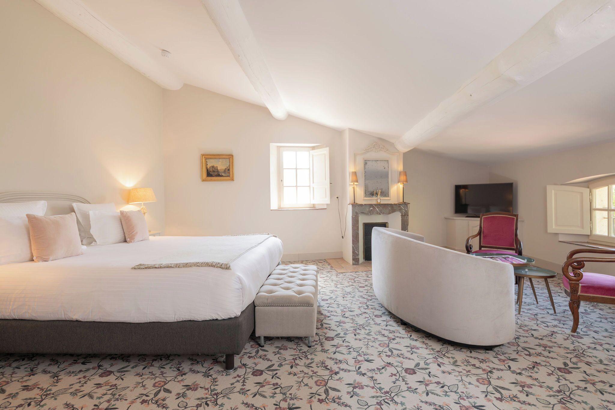 A bedroom with a large bed, a chair, a small table, a fireplace, and a television.
