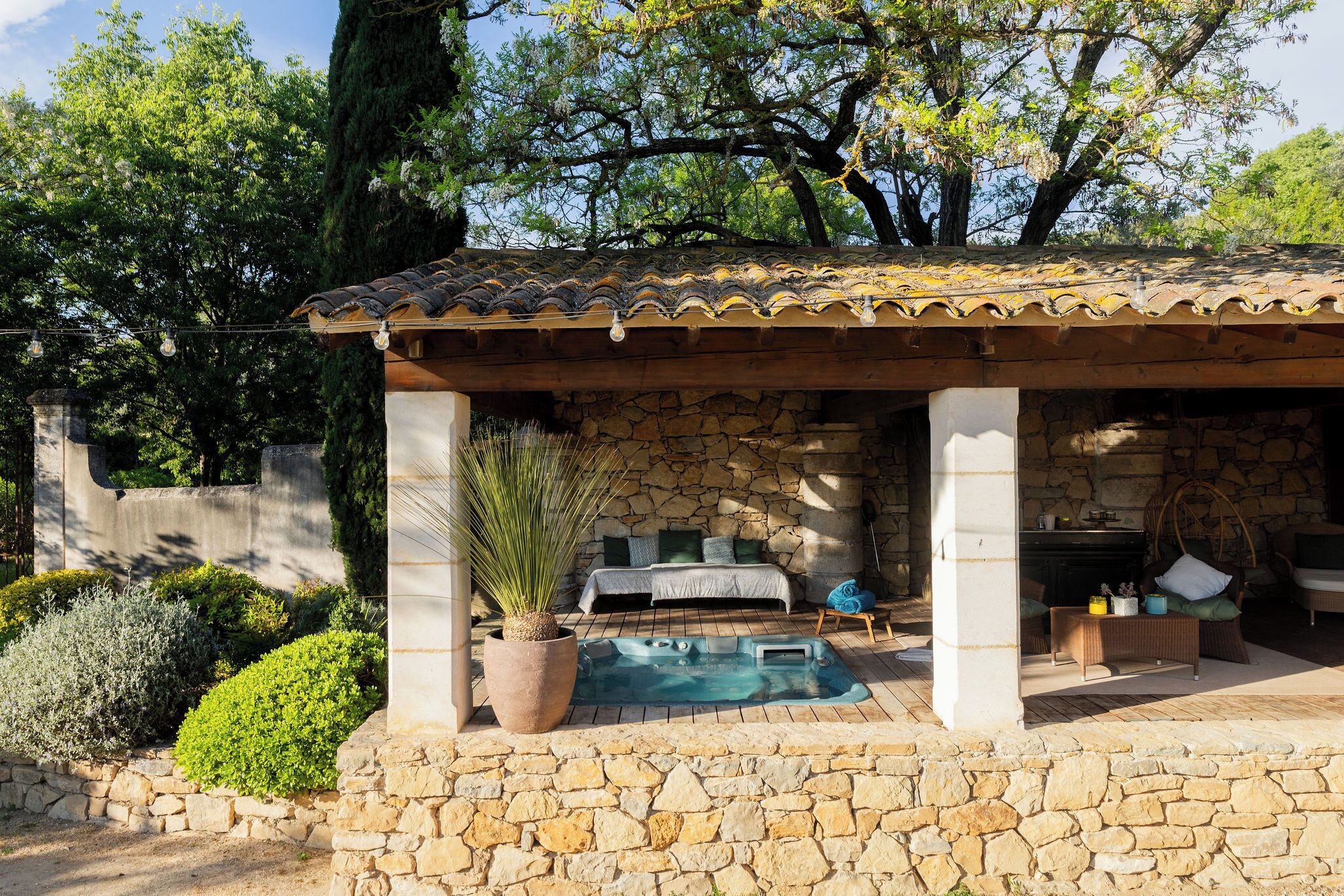 A stone-walled outdoor hot tub area with a bed and seating, surrounded by greenery and trees.