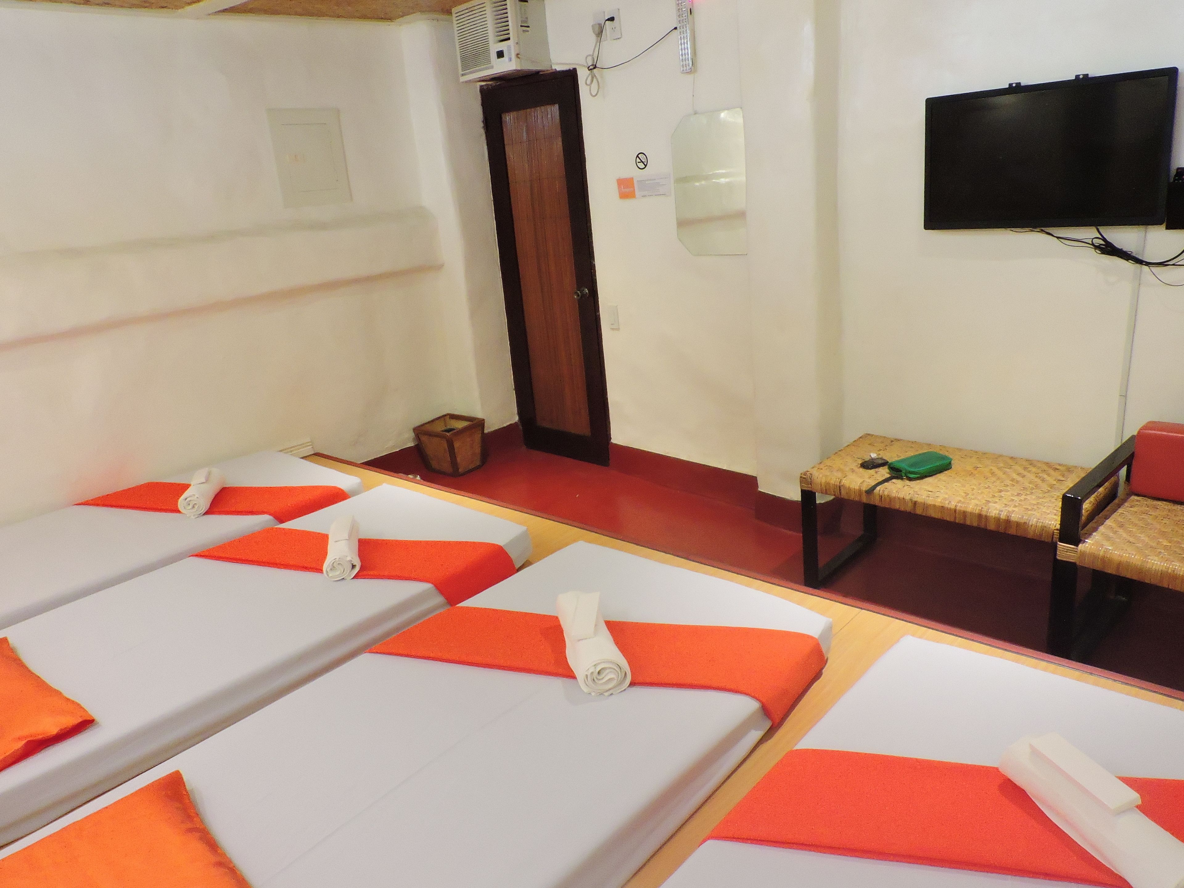 A room with four beds, each with white sheets and orange headboards, a wooden bench, a television mounted on the wall, and a small wooden box on the floor.