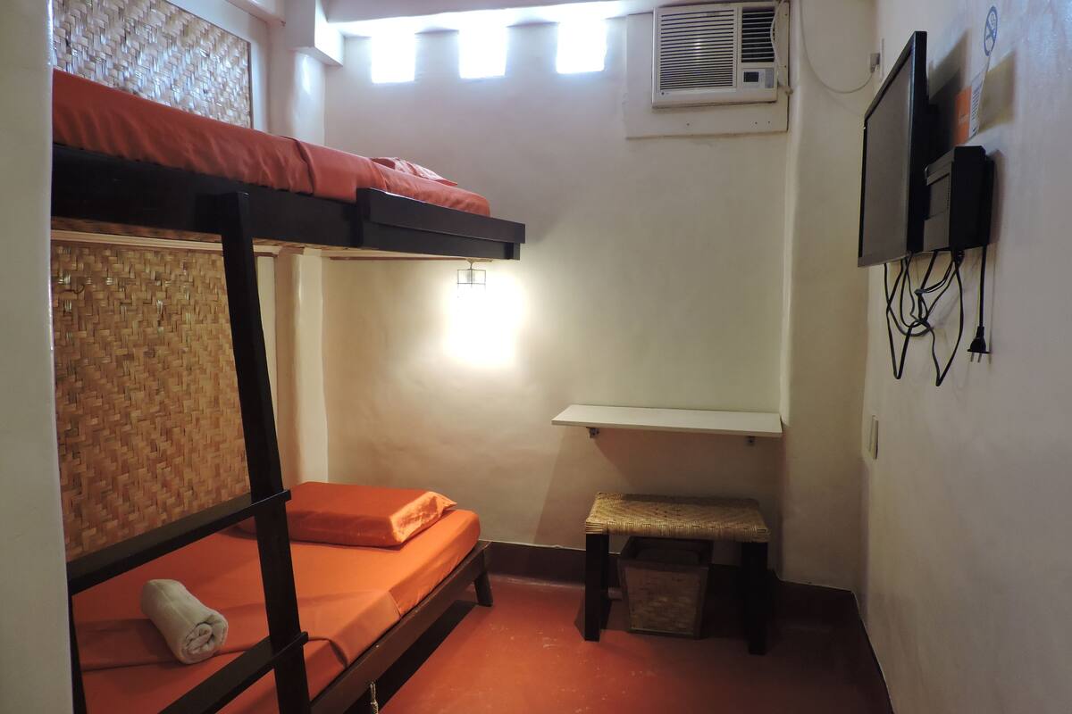 A small, compact room with a bunk bed, a single bed, a small bench, a wall-mounted TV, and an air conditioning unit.