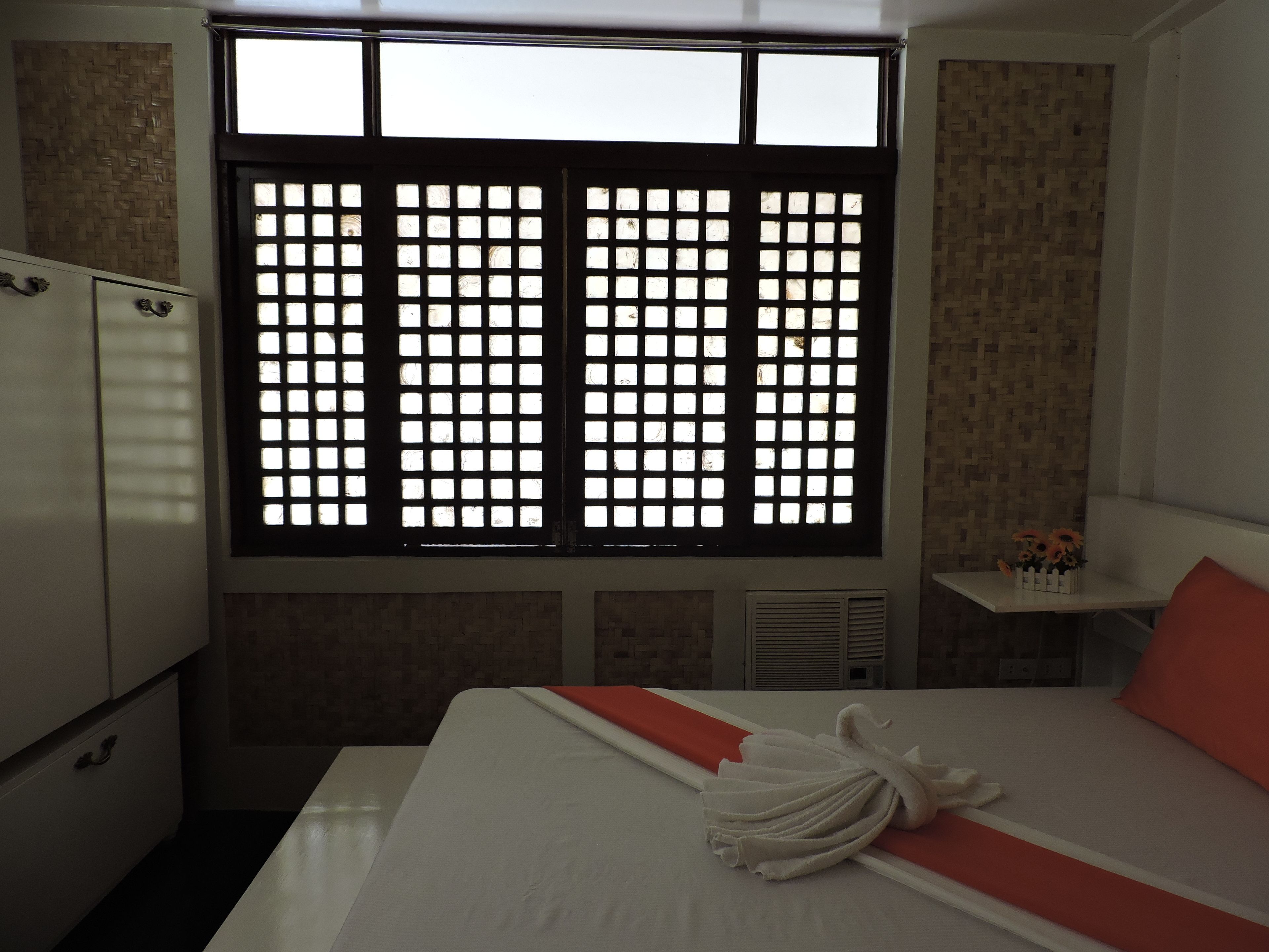 A hotel room with a bed, a window with a grid pattern, a white wardrobe, and a small shelf with flowers.