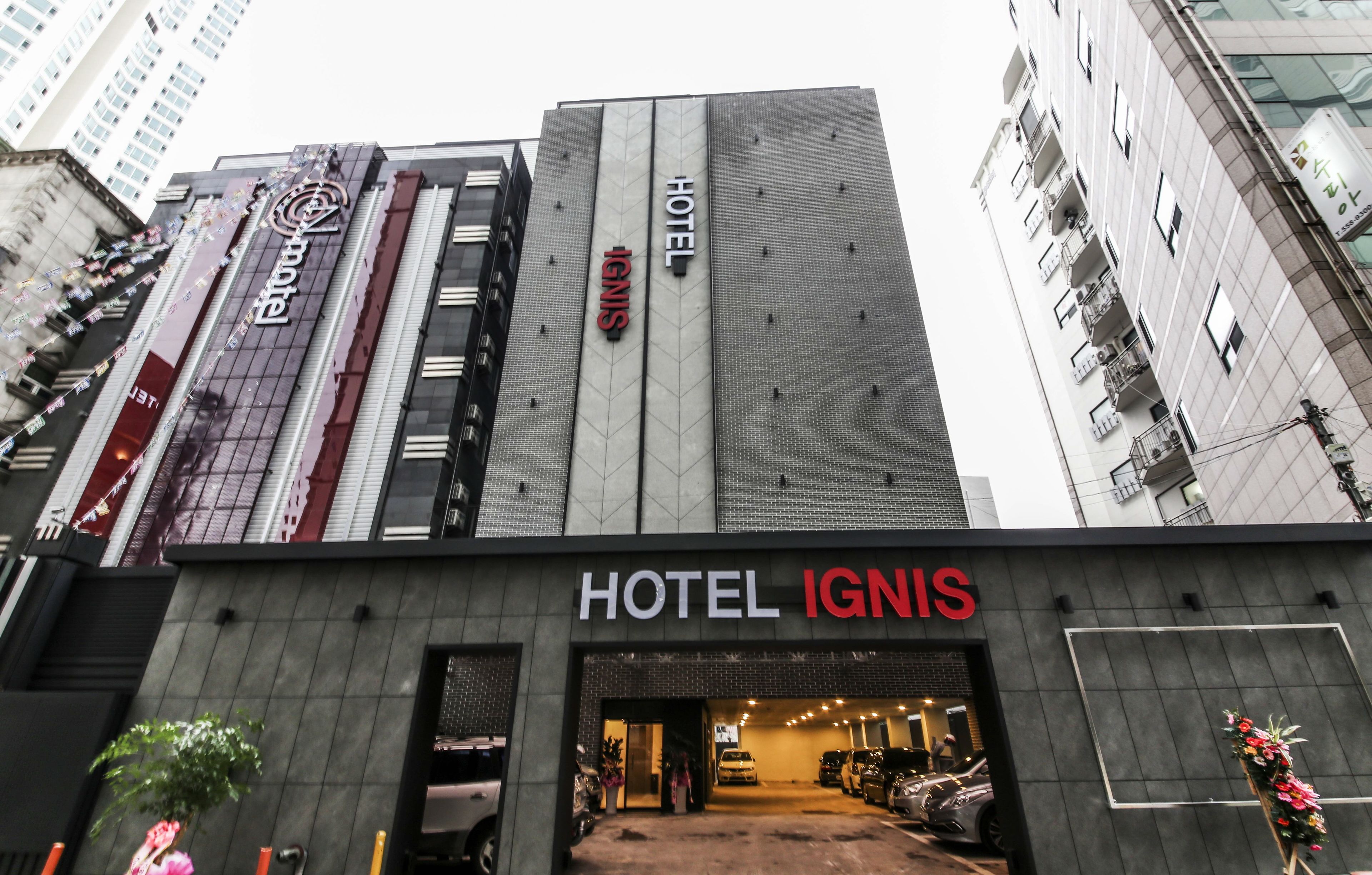 A hotel entrance with a modern facade, signage for "HOTEL IGNIS," and a parking area visible inside.