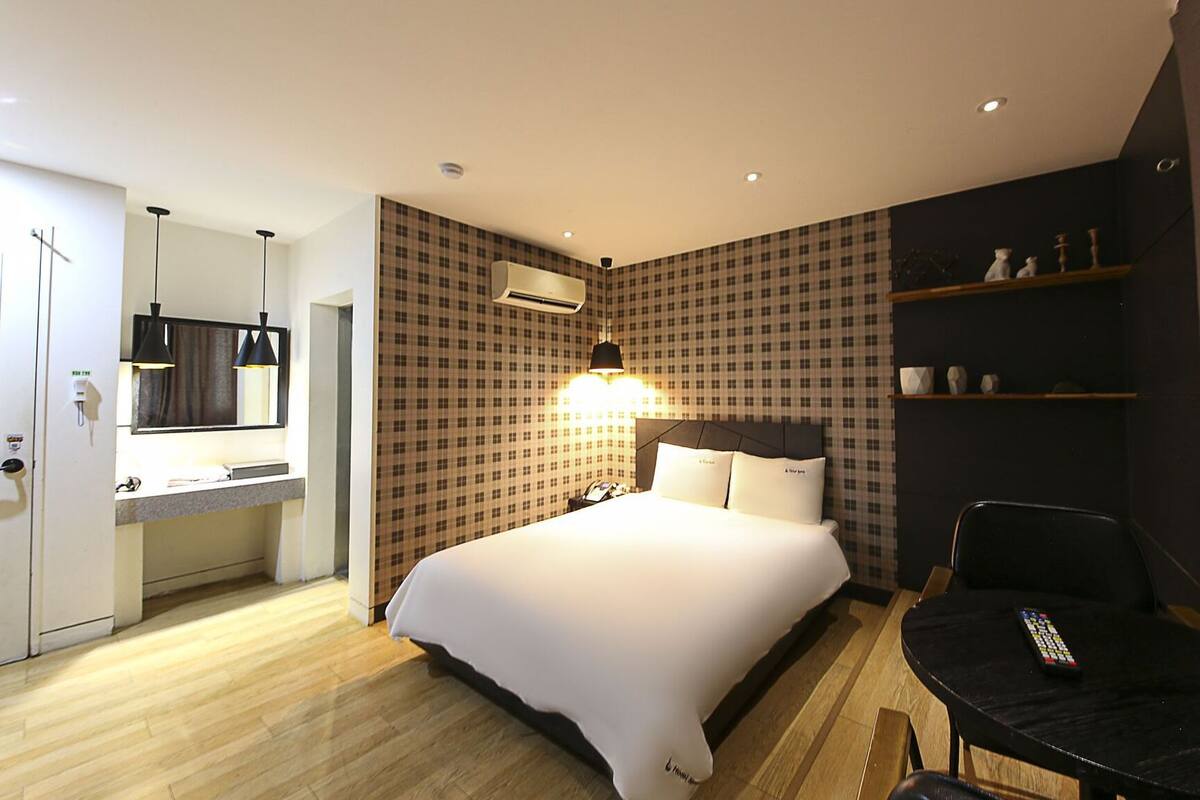 Une chambre d’hôtel comprenant un lit, un bureau avec une chaise, un miroir, une étagère avec des objets décoratifs et un climatiseur fixé au mur.