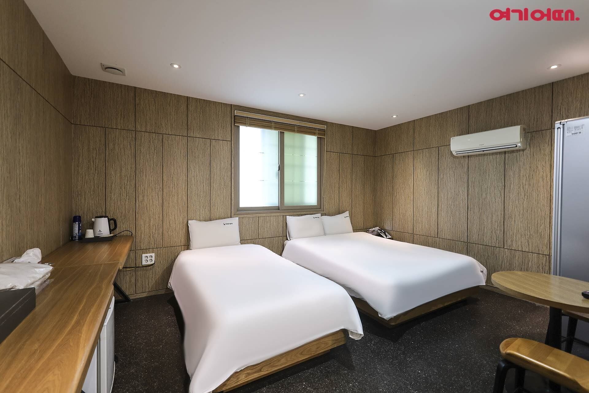 A hotel room with two beds, a wooden panel wall, a window with blinds, an air conditioning unit, and a small table.