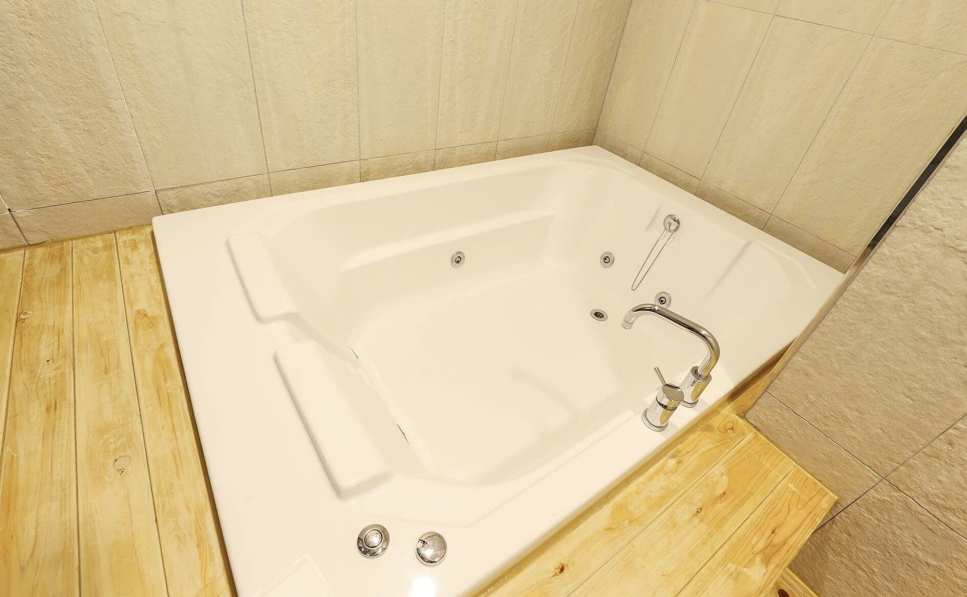 Eine weiße Badewanne mit Duschkopf und zwei Wasserhähnen, eingebettet in einen Raum mit Holzboden und beigen Fliesenwänden.