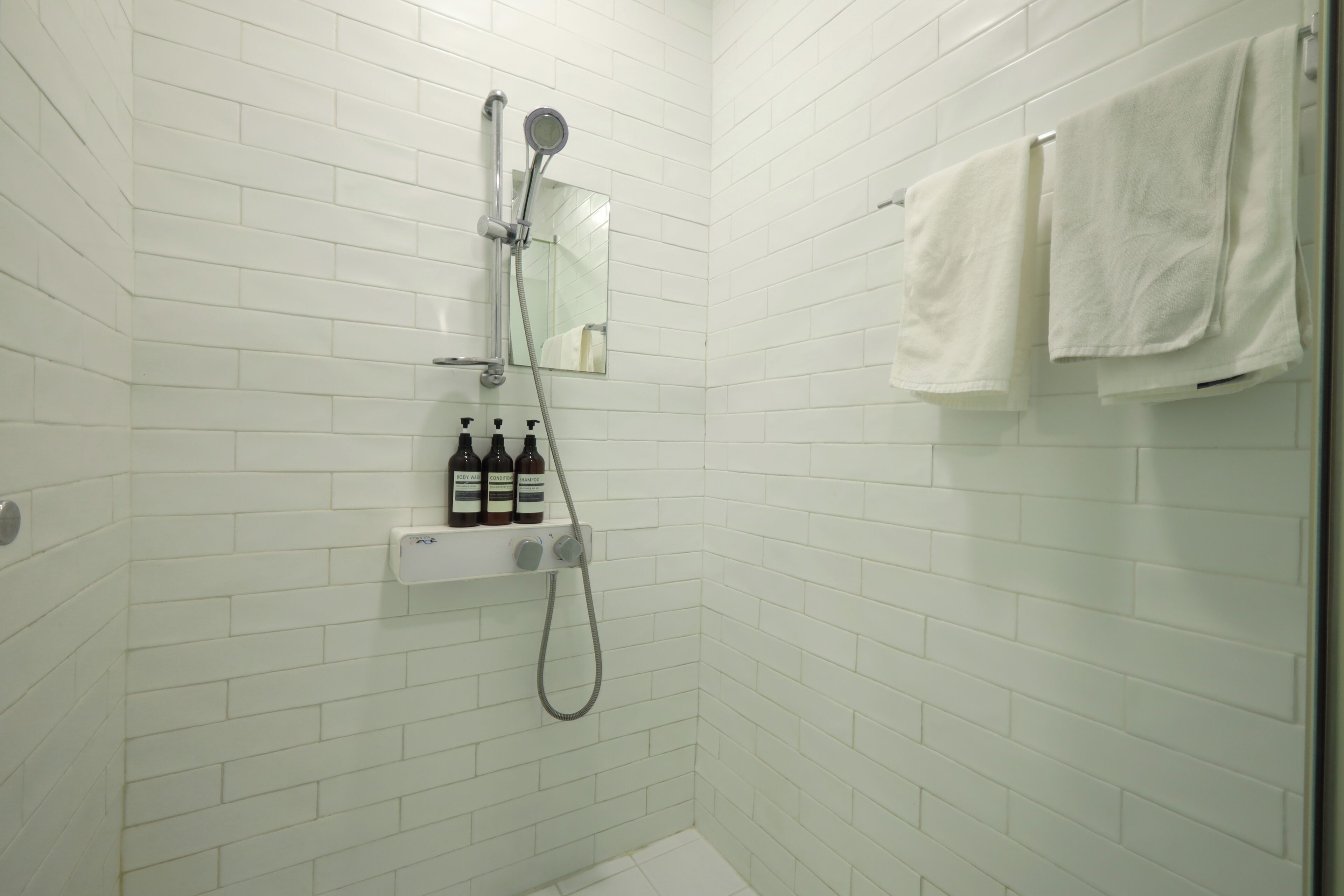 Ducha con azulejos blancos, cabezal de ducha de mano y repisa con botellas.