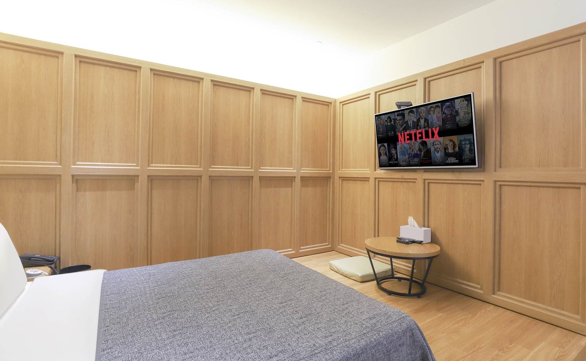 A hotel room with a bed, a TV mounted on the wall, a small round table with a tissue box, and wooden paneling on the walls.