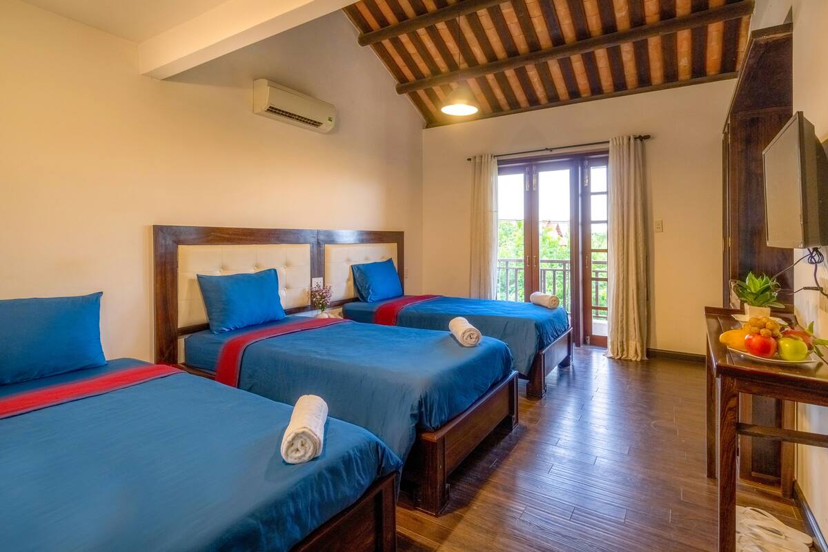 A hotel room with three beds, wooden flooring, a balcony with curtains, and a small table with fruits.