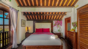 A bedroom with a wooden ceiling, a bed with red pillows, a nightstand, a dresser, and a window with a view of the countryside. - Flower’s House Hoi An (Da Nang)