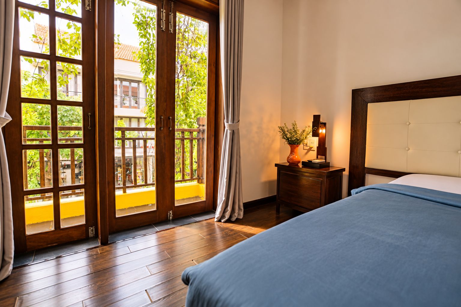 Habitación de hotel con piso de madera, cama con cubrecama azul, mesita de noche con un florero y una lámpara, y vista a un balcón exterior a través de una puerta de vidrio.