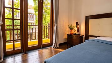 Habitación de hotel con piso de madera, cama con cubrecama azul, mesita de noche con un florero y una lámpara, y vista a un balcón exterior a través de una puerta de vidrio.